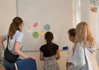 Vier Kinder und eine Betreuerin stehen vor einer Tafel mit bunten Aufklebern und Notizen.