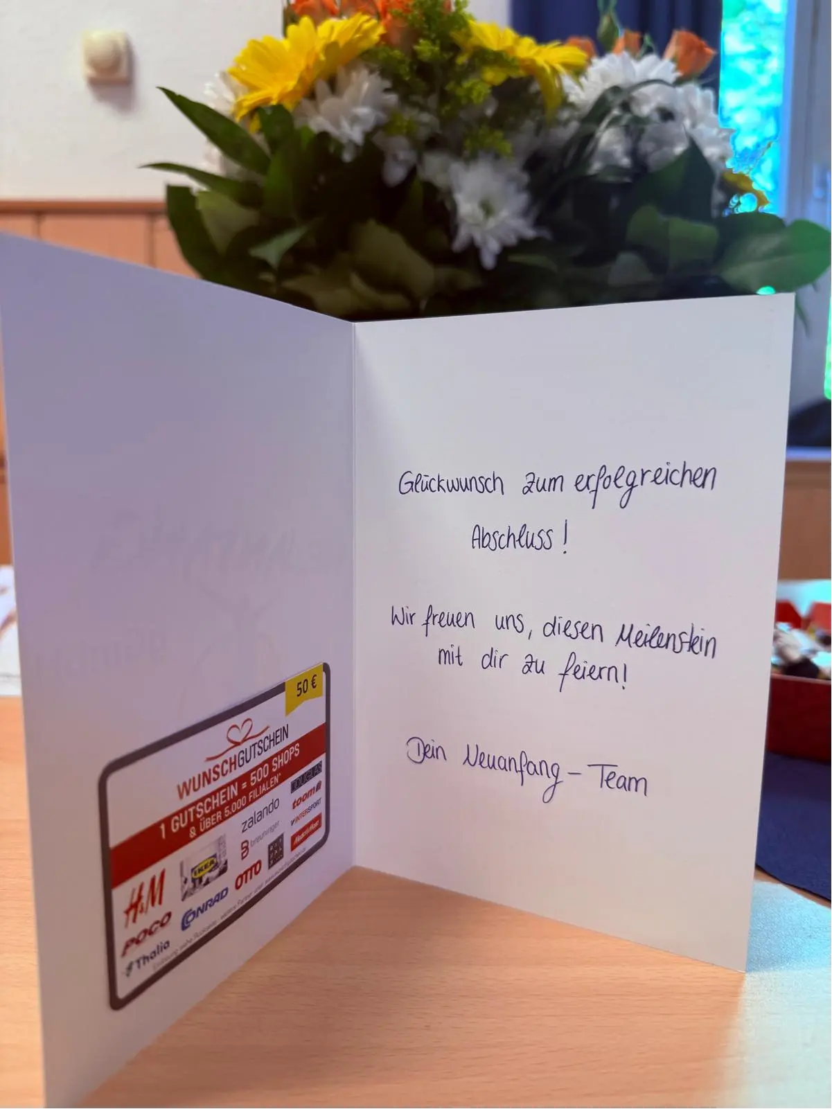 erfolgreiche-abschluesse-3 Eine Glückwunschkarte mit einem Gutschein für verschiedene Geschäfte und einem Blumengebinde im Hintergrund.