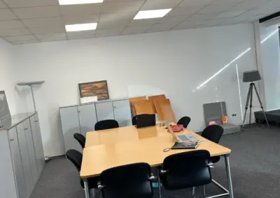Ein Besprechungsraum mit einem großen Tisch, Stühlen und einem Regal im Hintergrund. Helle Deckenbeleuchtung und neutraler Teppichboden.