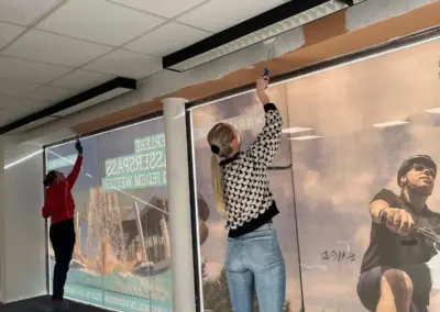 Zwei Personen, die an einer Wand mit großen Plakaten arbeiten, während sie die oberen Ränder mit einem Werkzeug anpassen.