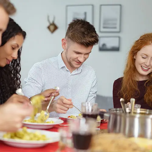 Mehrere junge Erwachsene essen gemeinsam an einem gedeckten Tisch und unterhalten sich in einem hellen Wohnraum mit freundlicher Atmosphäre.