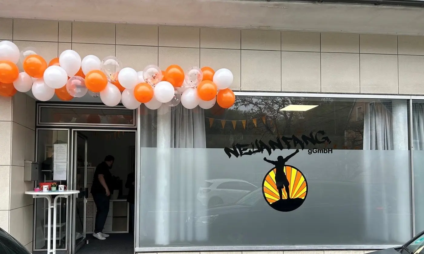 Vor dem Eingang der Neuanfang gGmbH in Warstein hängen orange-weiße Ballons, während im Innenraum eine Person steht und auf der Scheibe das Logo zu sehen ist.