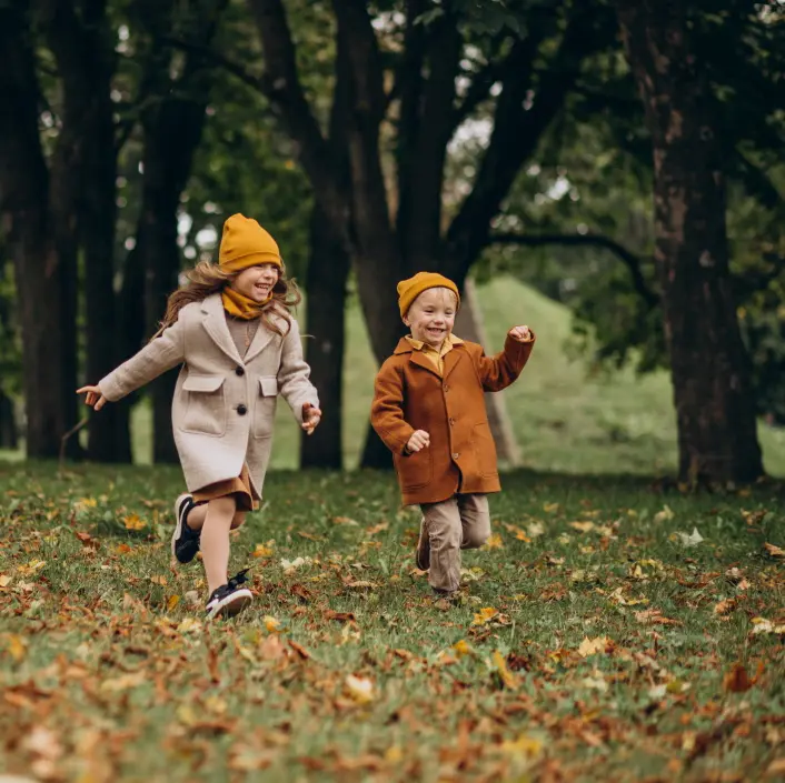Zwei Kinder laufen lachend durch einen herbstlichen Park, beide mit gelben Mützen und warmen Mänteln, während bunte Blätter den Boden bedecken.